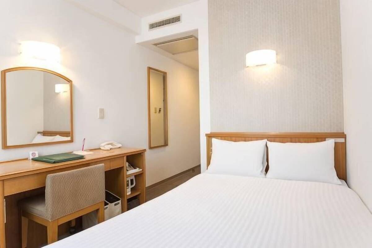 A hotel room with a bed, a desk with a chair, a mirror, and two wall-mounted lights.