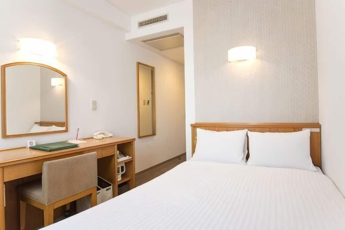 A hotel room with a bed, a desk with a chair, a mirror, and two wall-mounted lights.