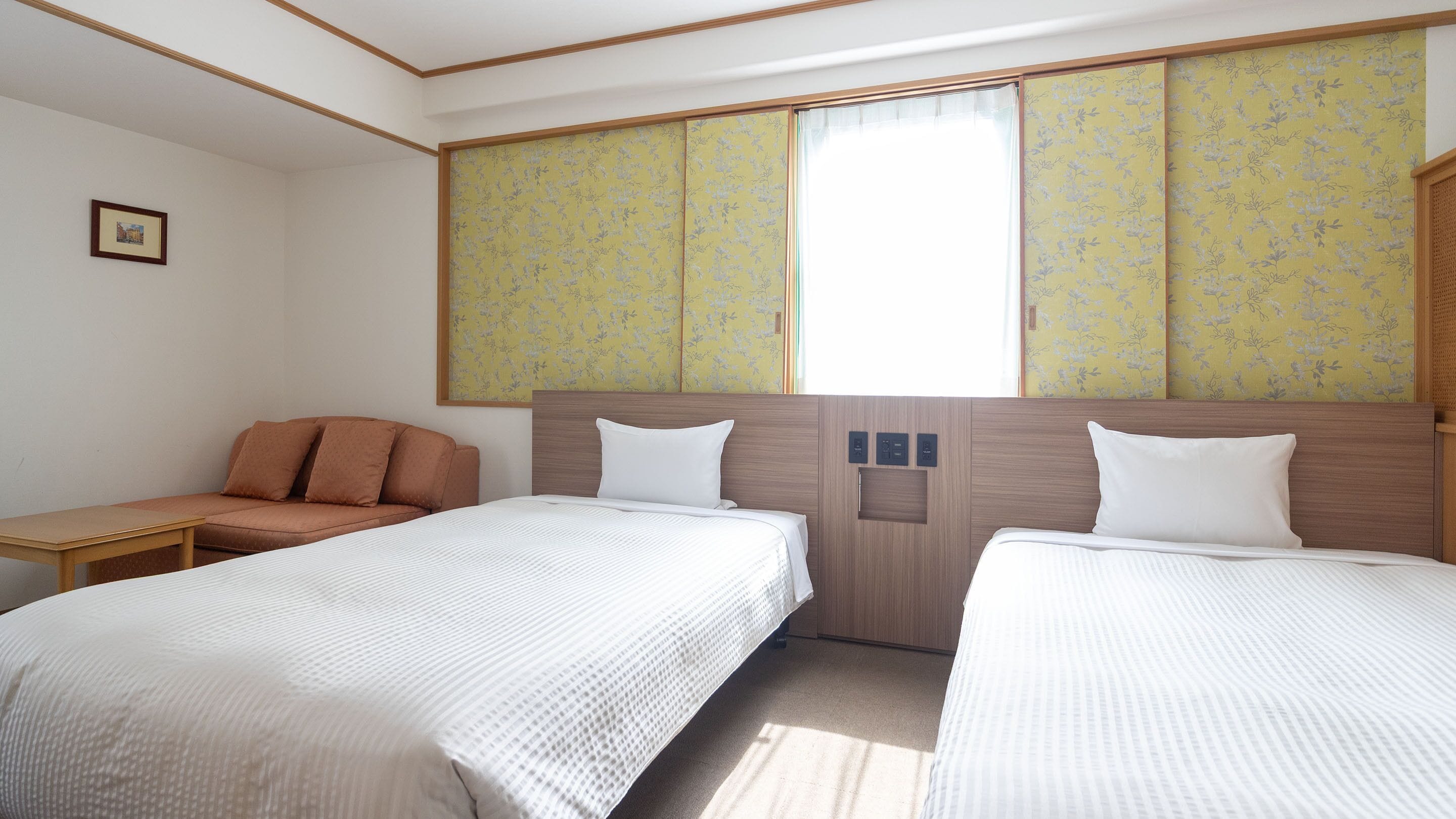 A hotel room with two beds, a sofa, a small table, and a window with floral wallpaper.