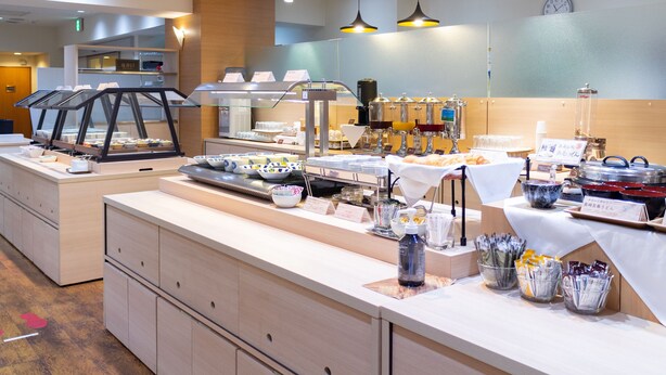 A breakfast buffet area with various food items and beverages, a coffee machine, and a clock on the wall.