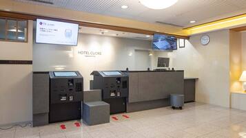 Zona de recepción de hotel con quioscos de autoservicio, un reloj y una pantalla digital que muestra información.