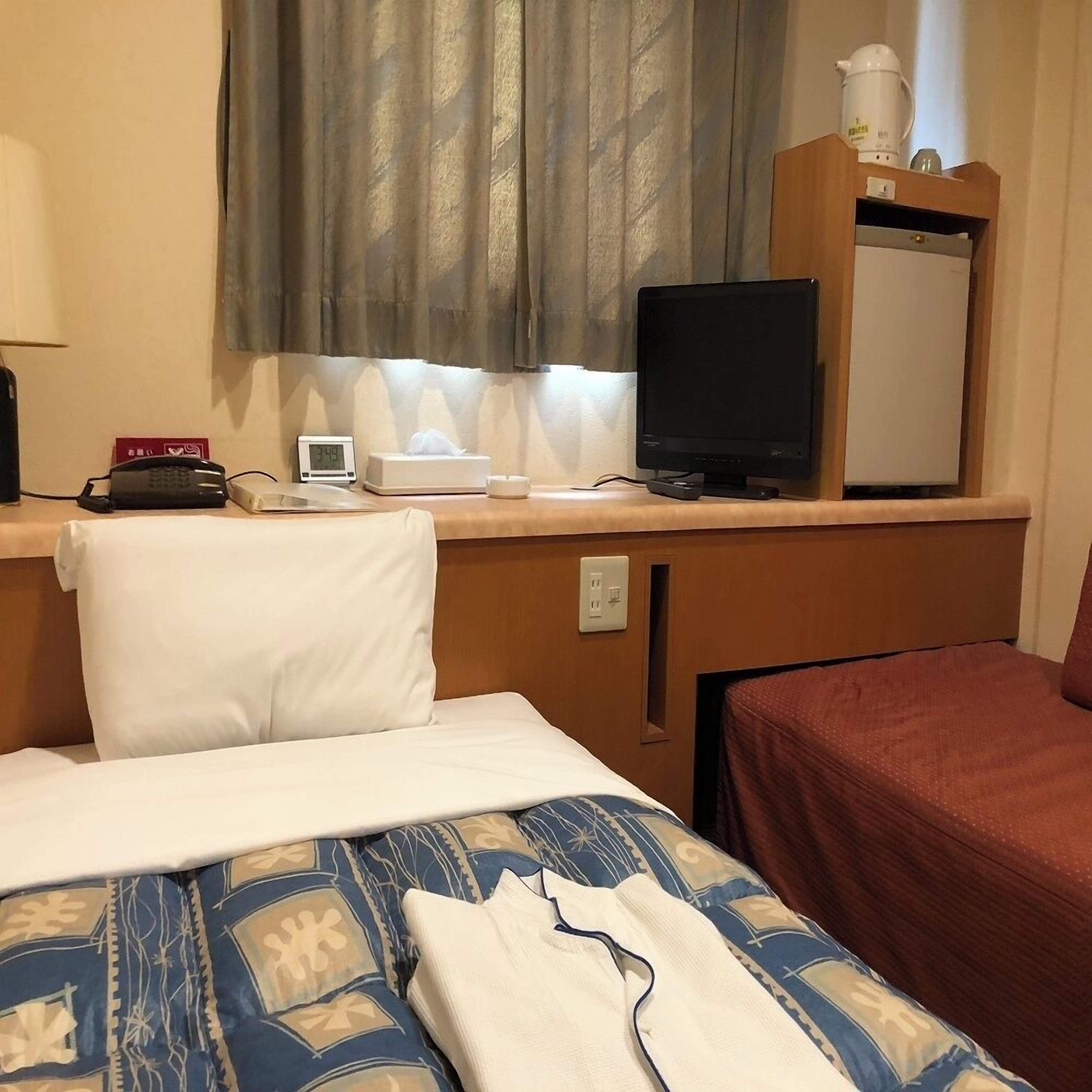 A hotel room with a bed, a desk with a computer, a telephone, a clock, and a window with curtains.