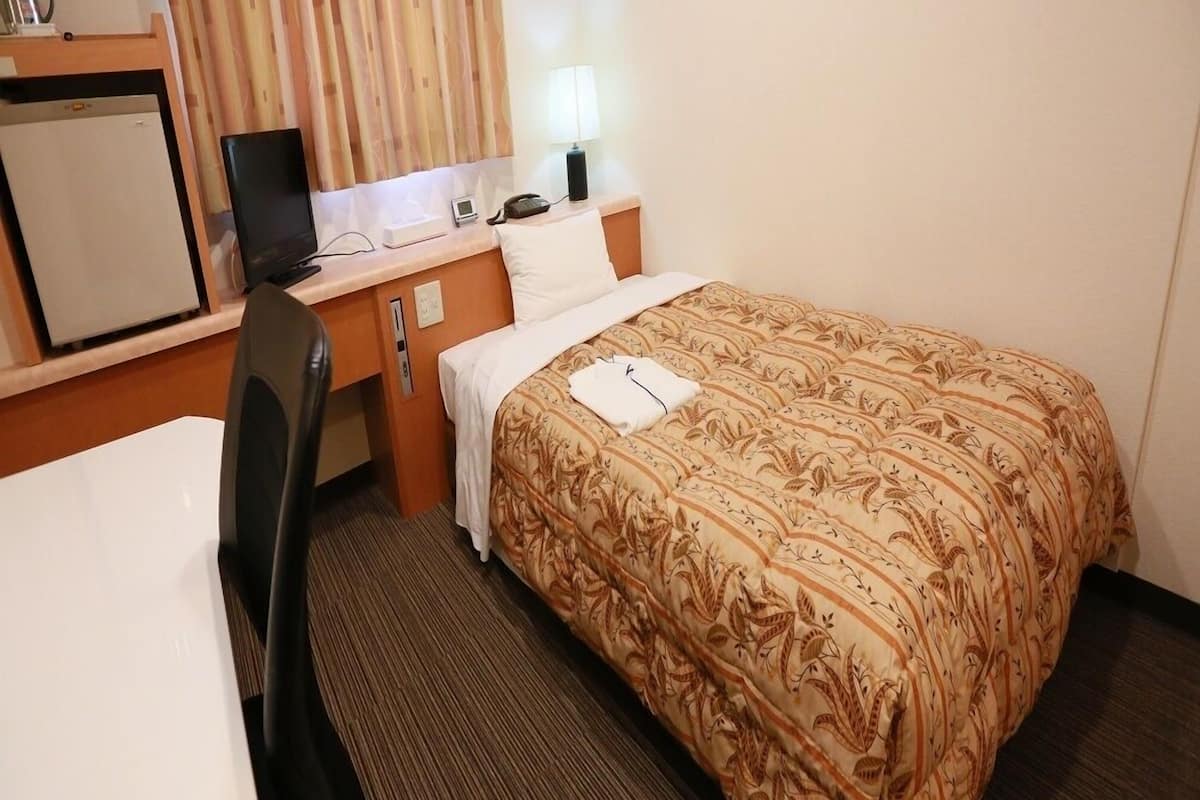 Habitación de hotel con cama, escritorio con computadora, silla, refrigerador pequeño y ventana con cortinas.