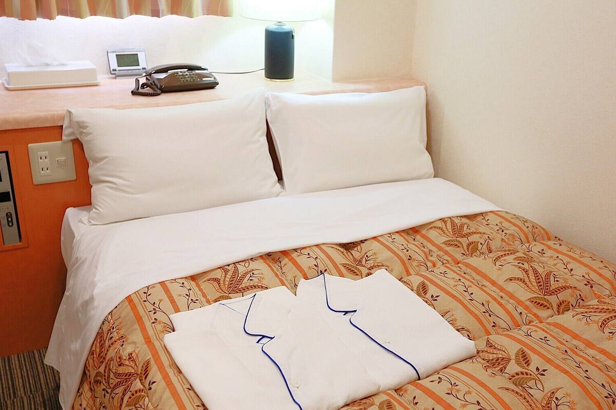 Una habitación de hotel con una cama, almohadas, una mesita de noche con teléfono, lámpara y toma de corriente.