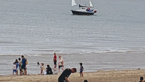 Une scène de plage avec un voilier sur l’eau et des personnes sur le rivage.