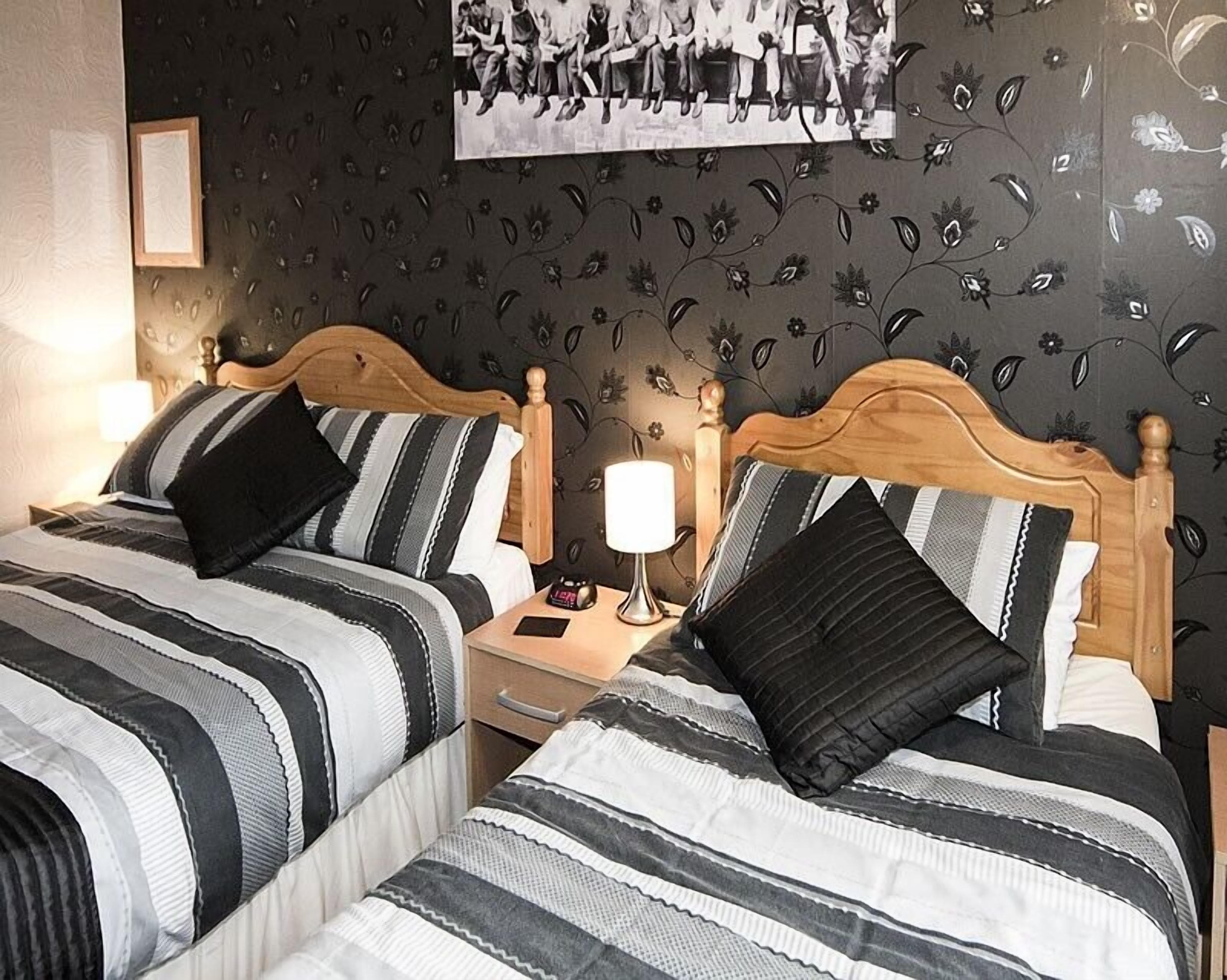 Une chambre d’hôtel avec deux lits, chacun doté de draps à rayures et d’une tête de lit en bois. On aperçoit une table de chevet avec une lampe, ainsi qu’une petite table sur laquelle se trouvent un téléphone et une horloge.