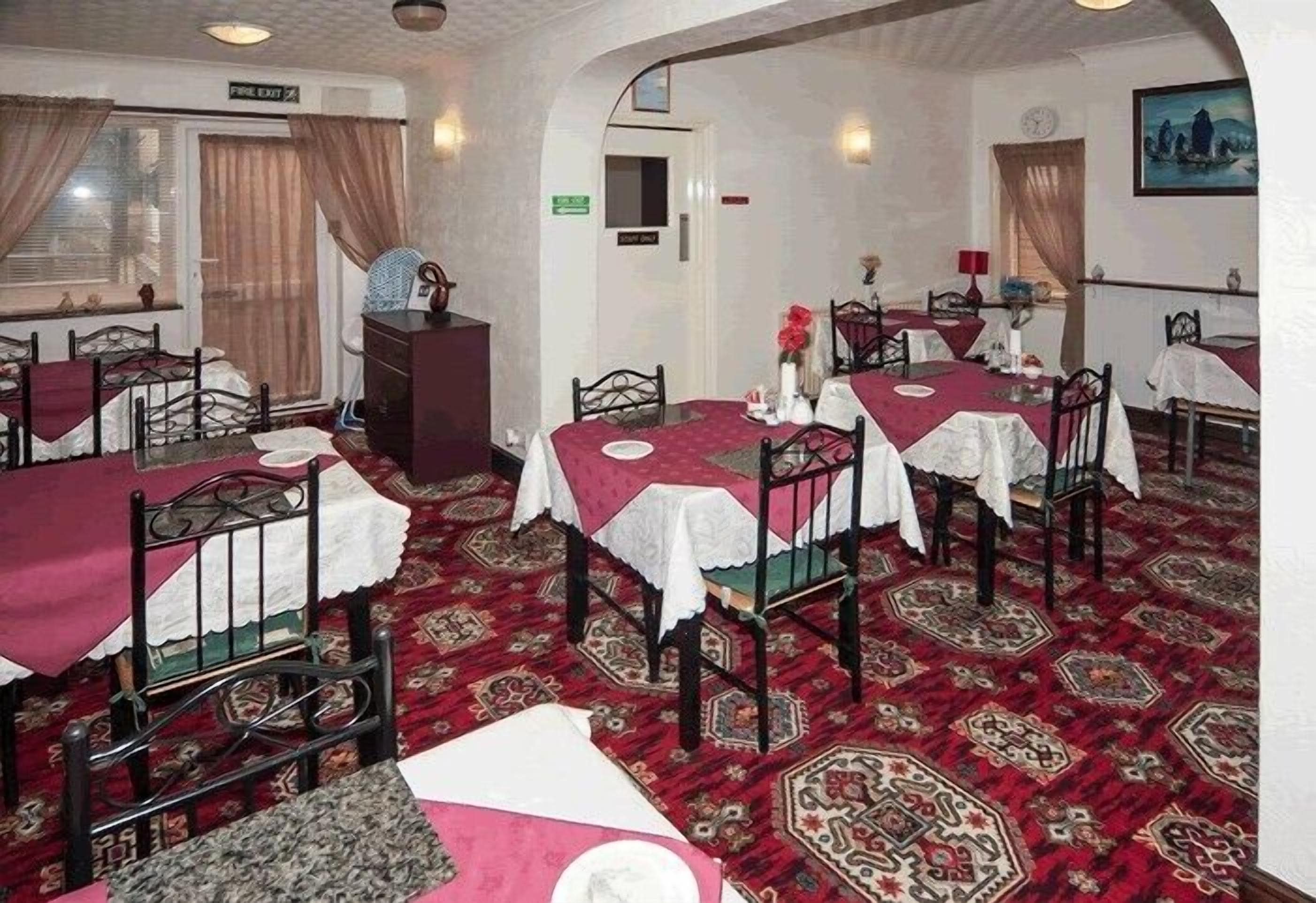 Un comedor con mesas dispuestas para cuatro, cada una con mantel rojo y camino de mesa blanco. La habitación tiene alfombra estampada, sillas de madera y un pequeño armario de madera.