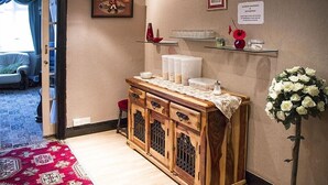 Un meuble en bois avec des portes en verre, un tapis rouge à motifs, un vase blanc rempli de fleurs et un panneau d’information sur les allergènes.