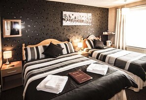 A hotel room with two beds, a dark patterned wall, a framed photo, a nightstand with a lamp, and a window with curtains.