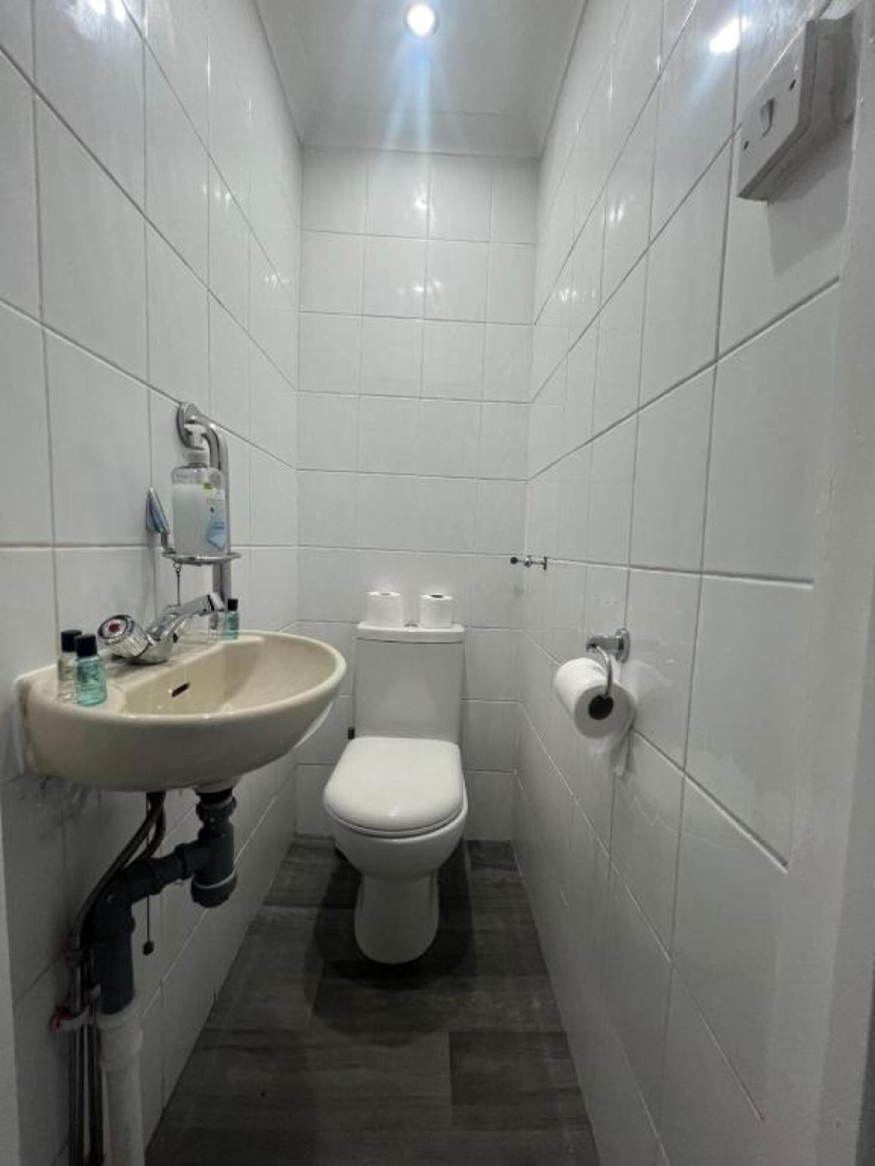 Un bagno piccolo e stretto, rivestito di piastrelle bianche, con un lavandino, un water e un portascirolettoni.