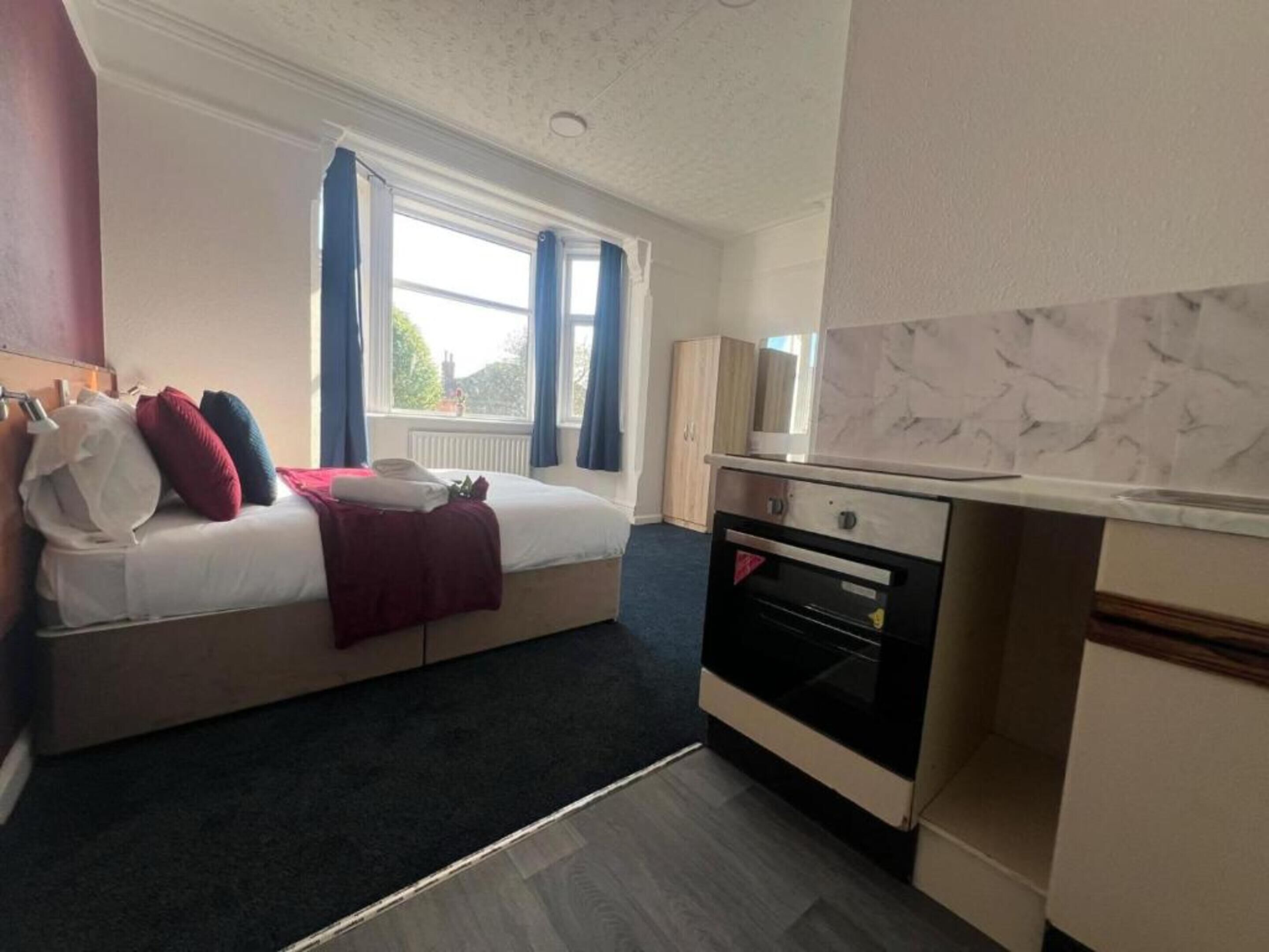 A compact hotel room with a bed, a small kitchenette, and a window with curtains.