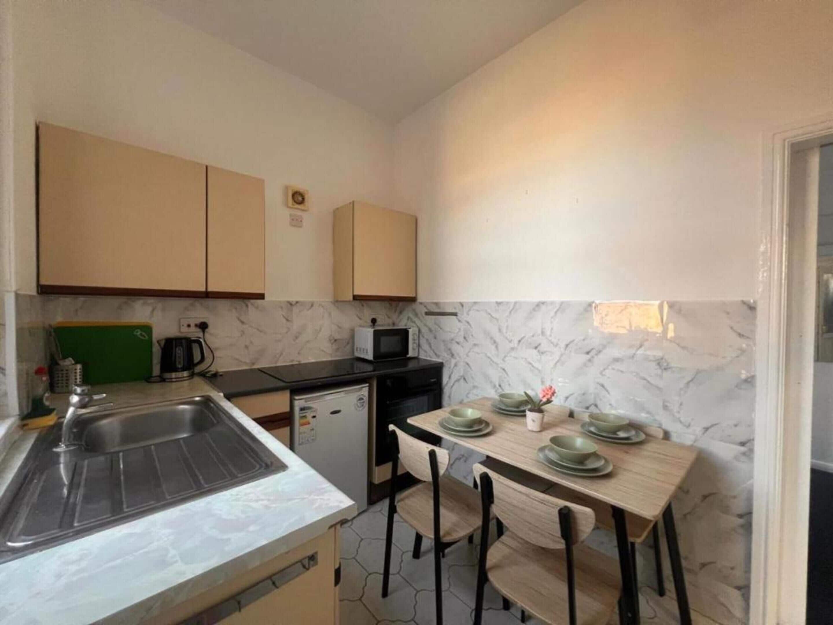 A compact kitchen with a sink, a microwave, and a small refrigerator. There is a dining table with chairs and a few plates.
