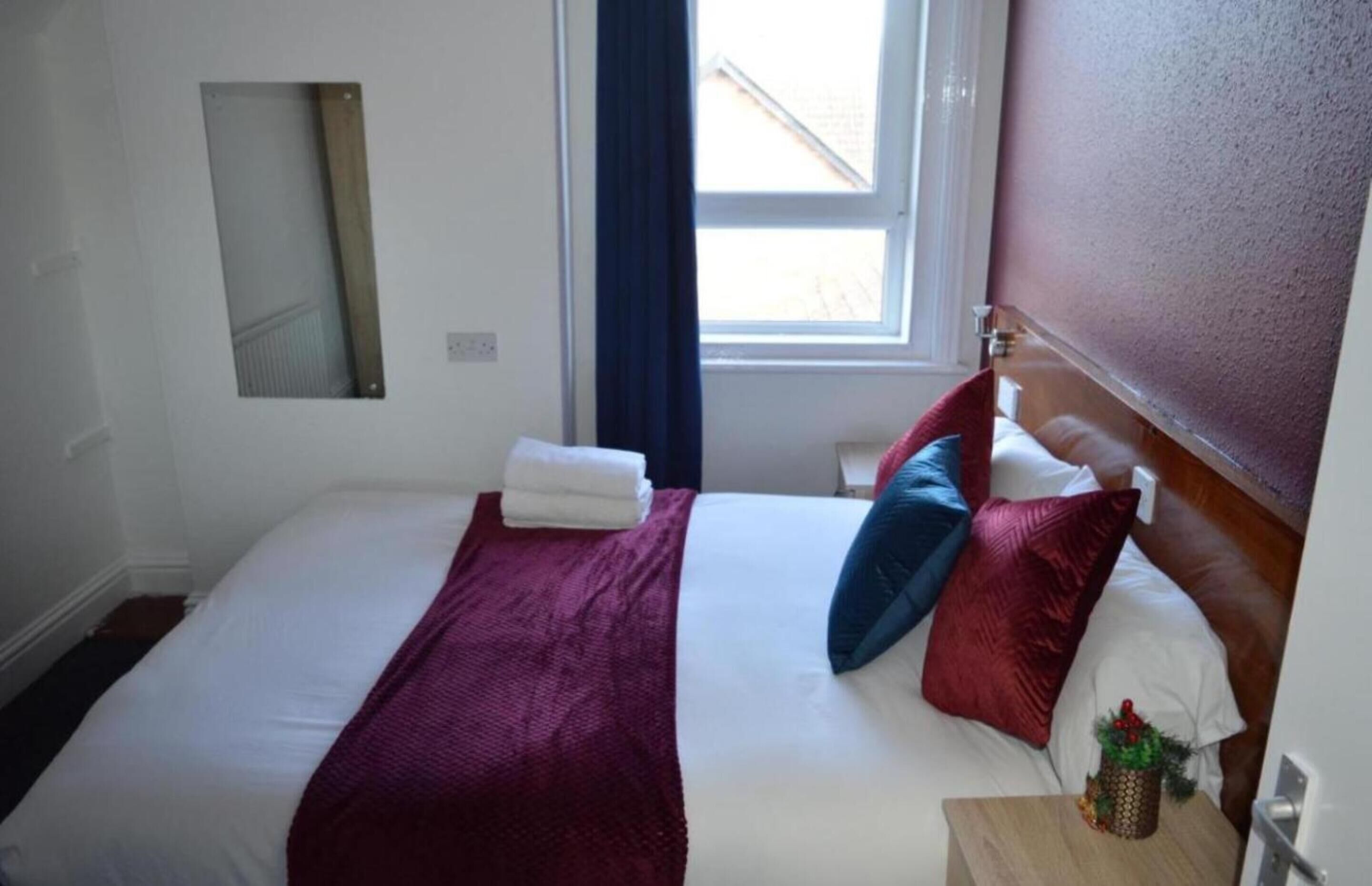 A neatly made bed with a red blanket and pillows, a small bedside table with a plant, and a window with a view of the outside.