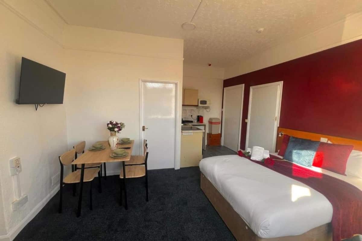 A compact hotel room with a single bed, a small kitchenette, a dining area with a table and chairs, and a TV mounted on the wall.