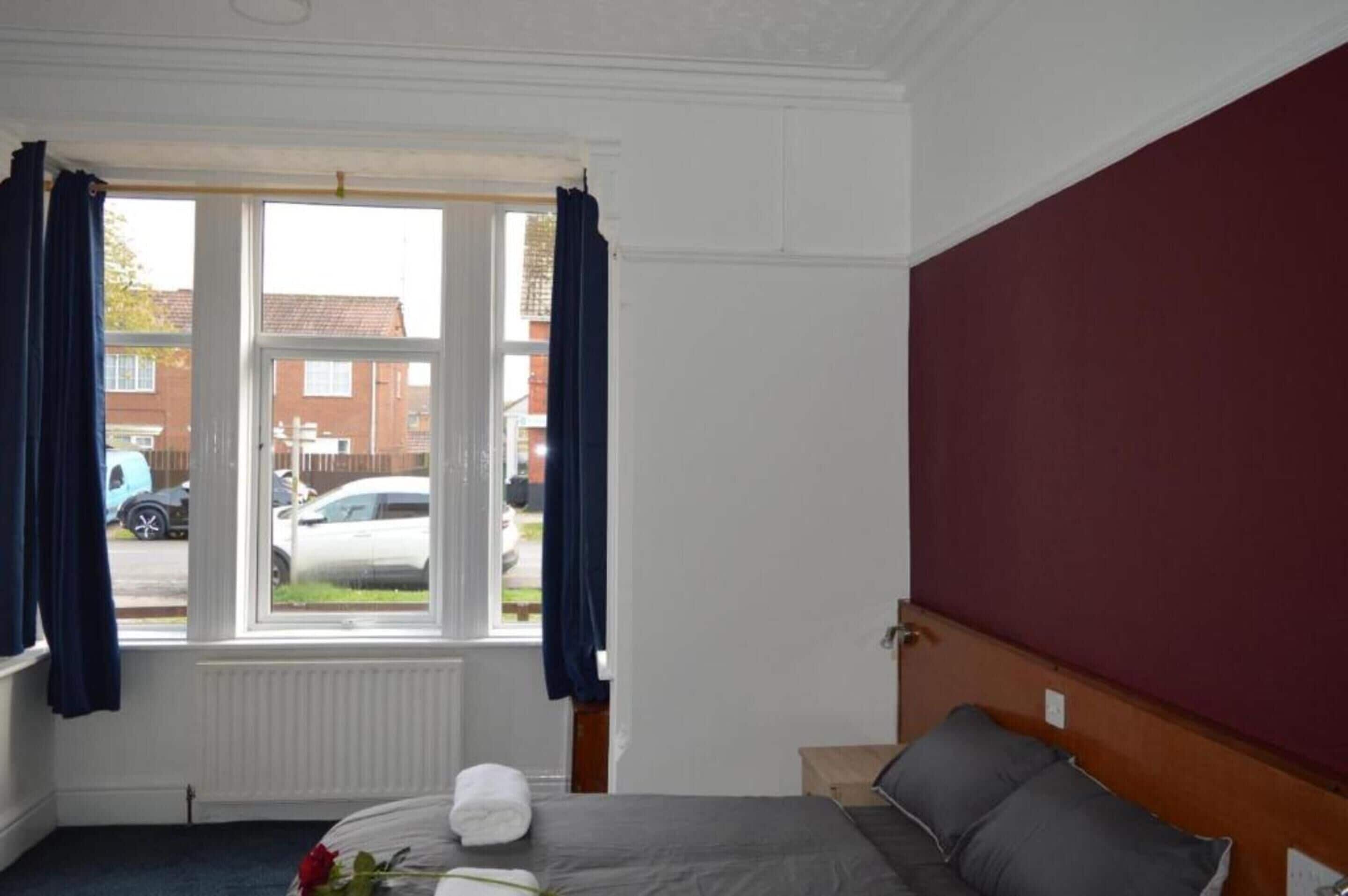 A bedroom with a bed, a window with curtains, a radiator, and a view of the street outside.