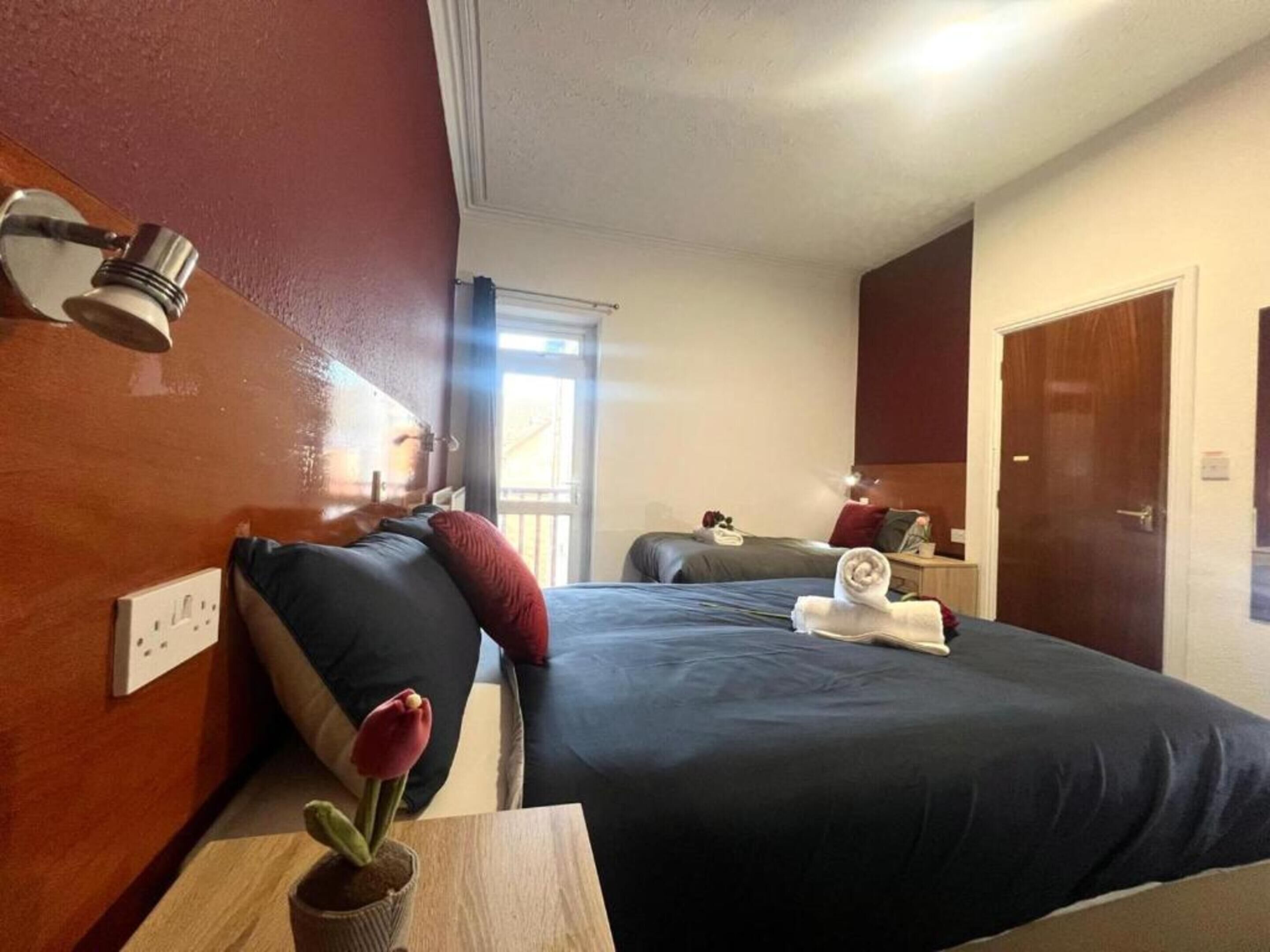 Une chambre d’hôtel avec deux lits, une tête de lit en bois, une table de chevet, une fenêtre avec des rideaux et une lampe fixée au mur.