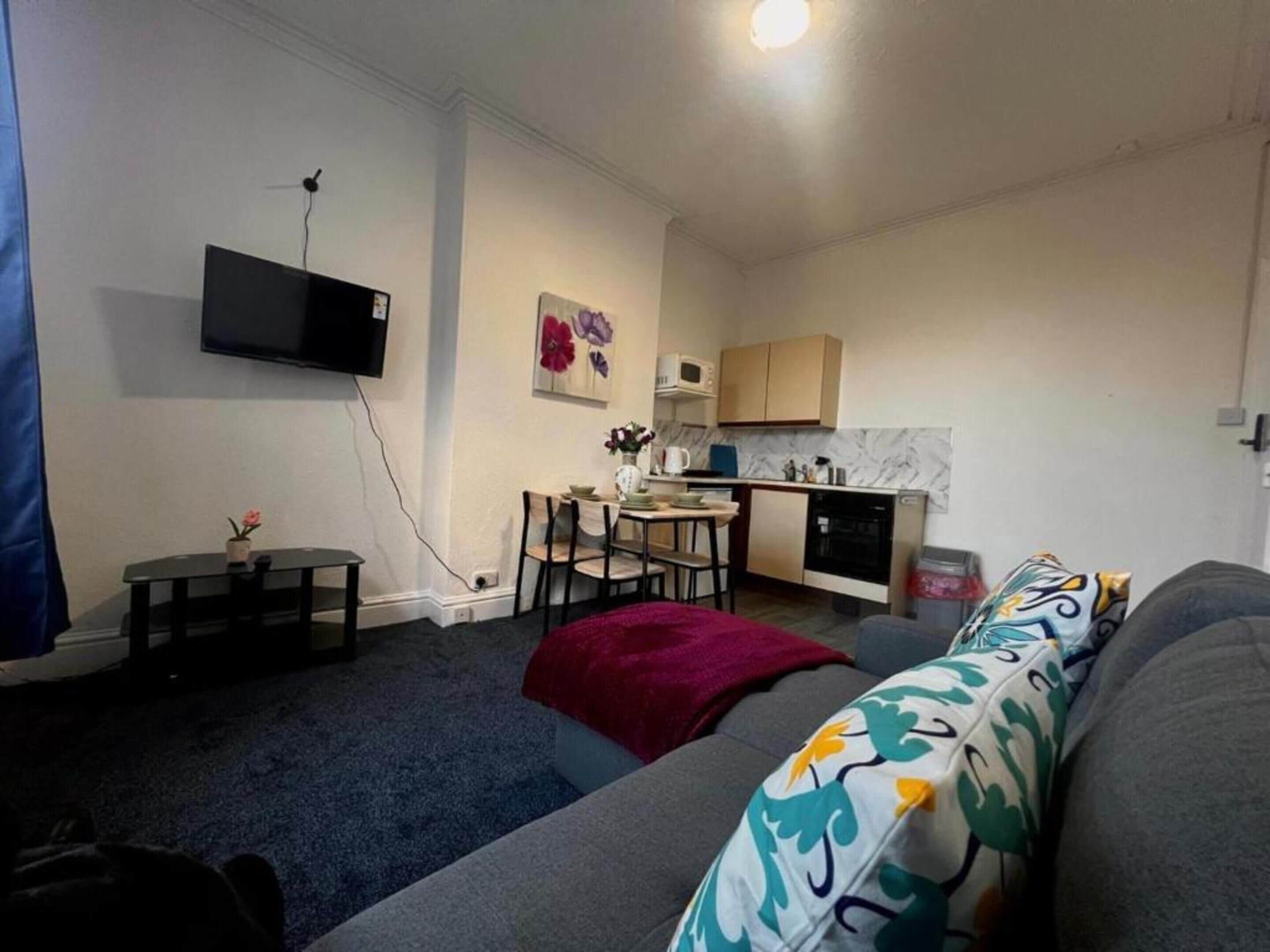 Un espacio habitable compacto con sofá, una mesa de comedor con sillas, una zona de cocina con armarios y un microondas, y un televisor de pantalla plana montado en la pared.