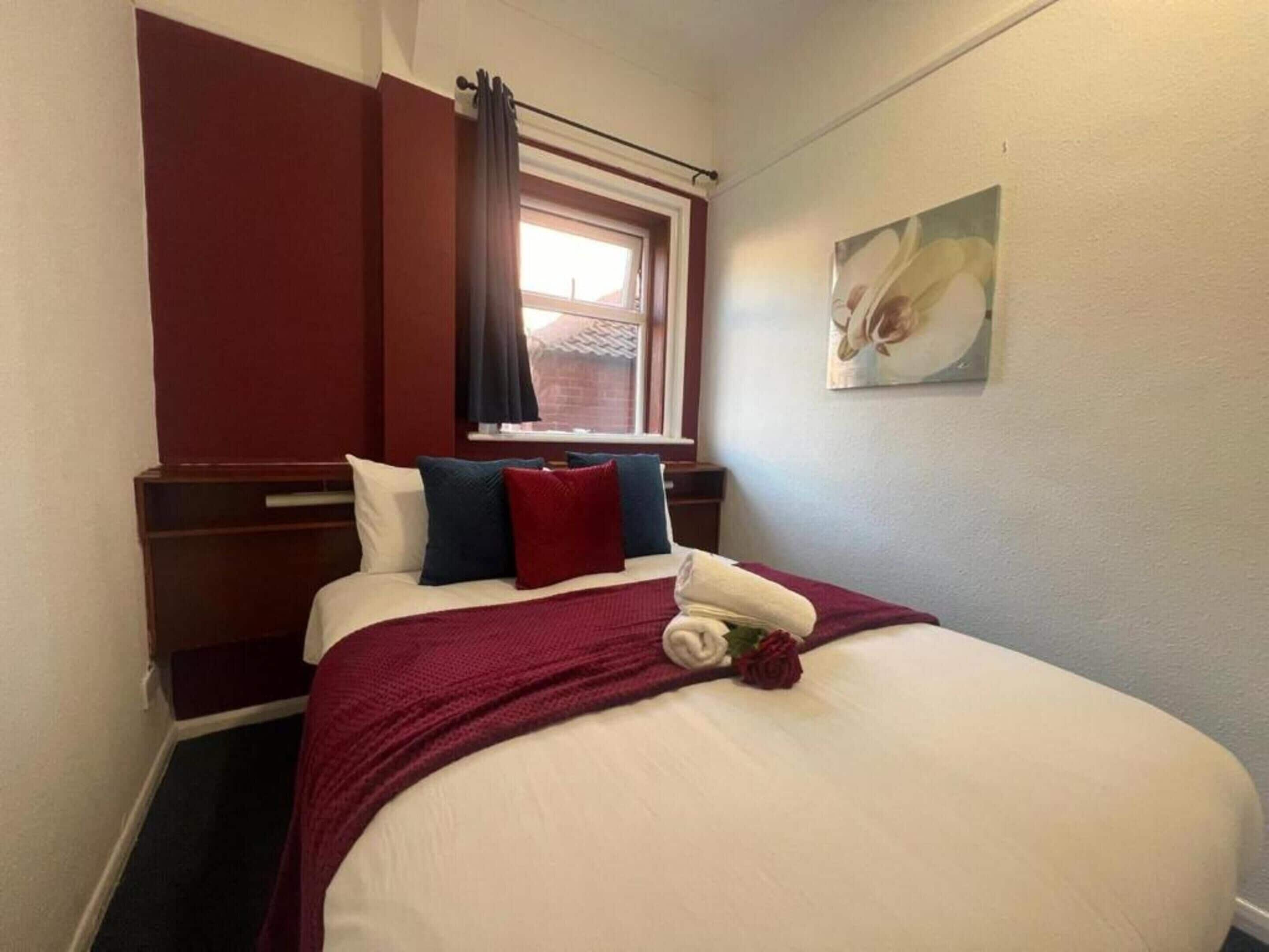A neatly made bed with a white comforter, red and blue pillows, and a red throw blanket. There is a window with curtains, a painting on the wall, and a wooden headboard.
