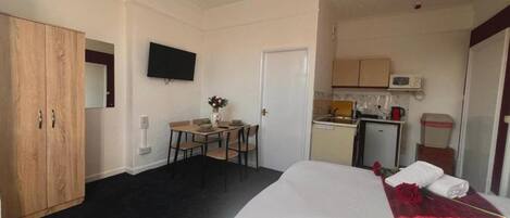A hotel room with a bed, a desk with a chair, a small kitchenette, and a wardrobe.