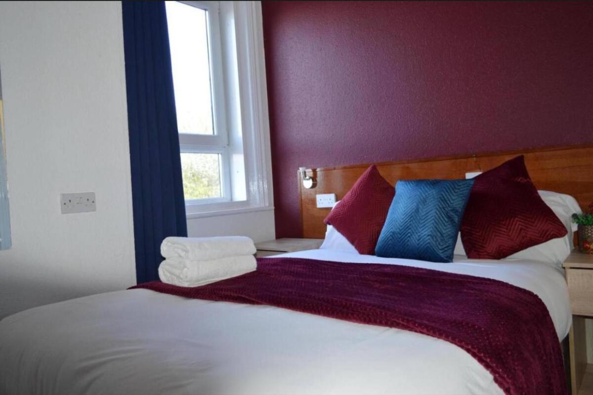A neatly made bed with white linens and a red throw blanket, a wooden headboard, and a nightstand with a lamp.
