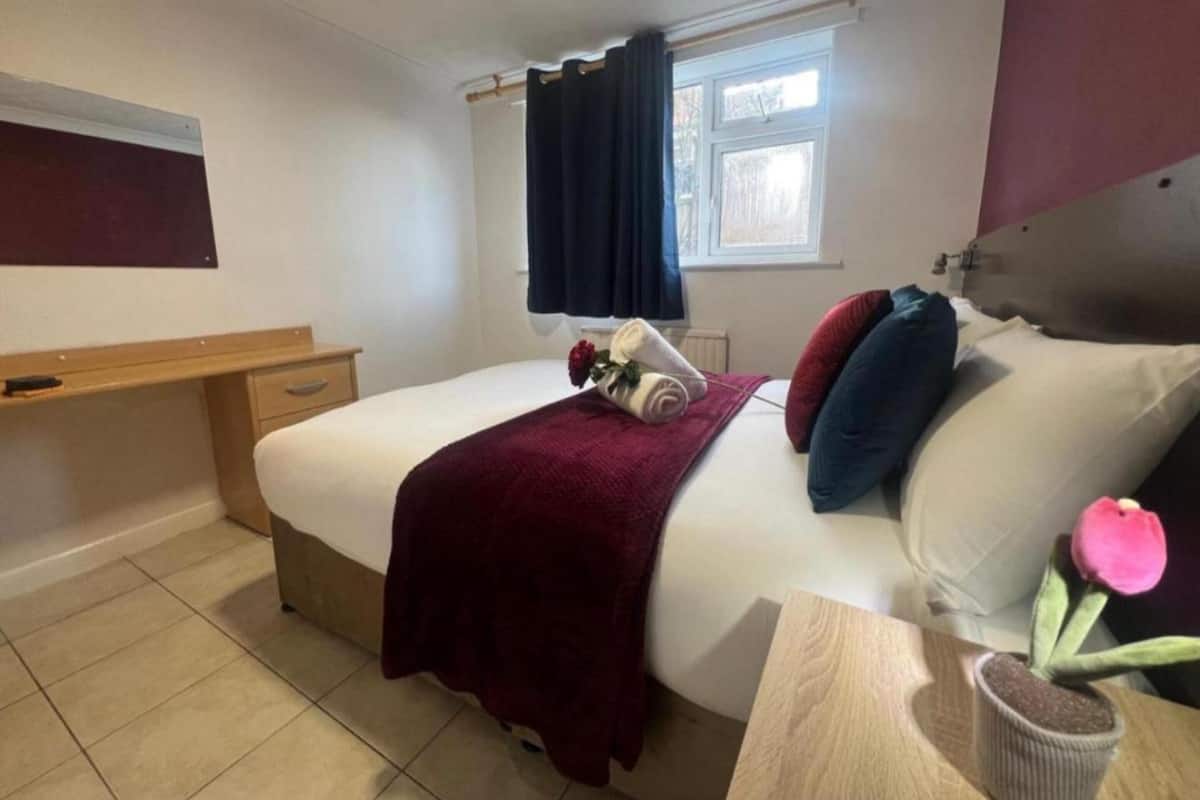 A neatly arranged bedroom with a bed, a bedside table with a vase of flowers, a window with curtains, and a small desk.
