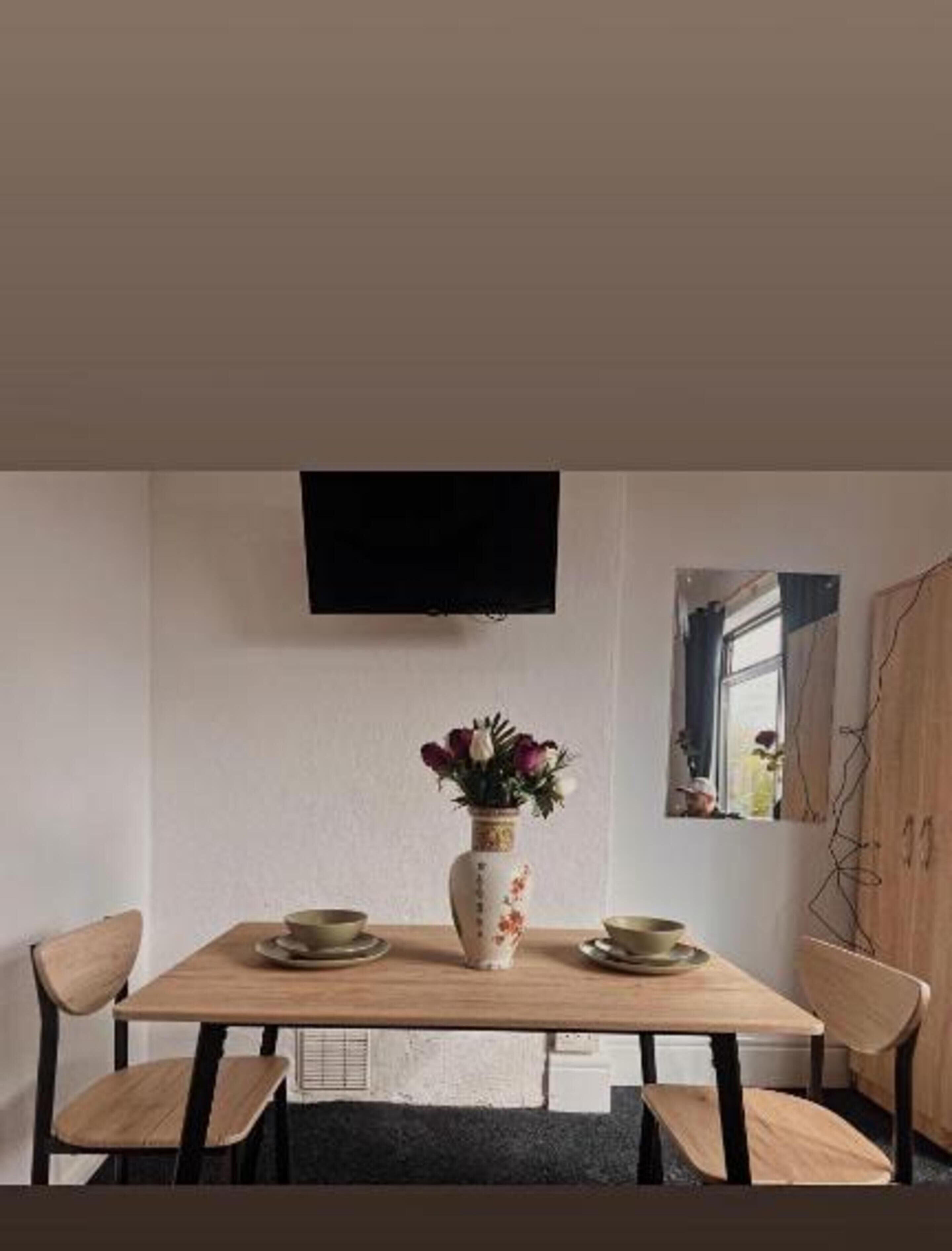 A dining area with a wooden table, two chairs, a vase with flowers, and a flat-screen TV mounted on the wall.