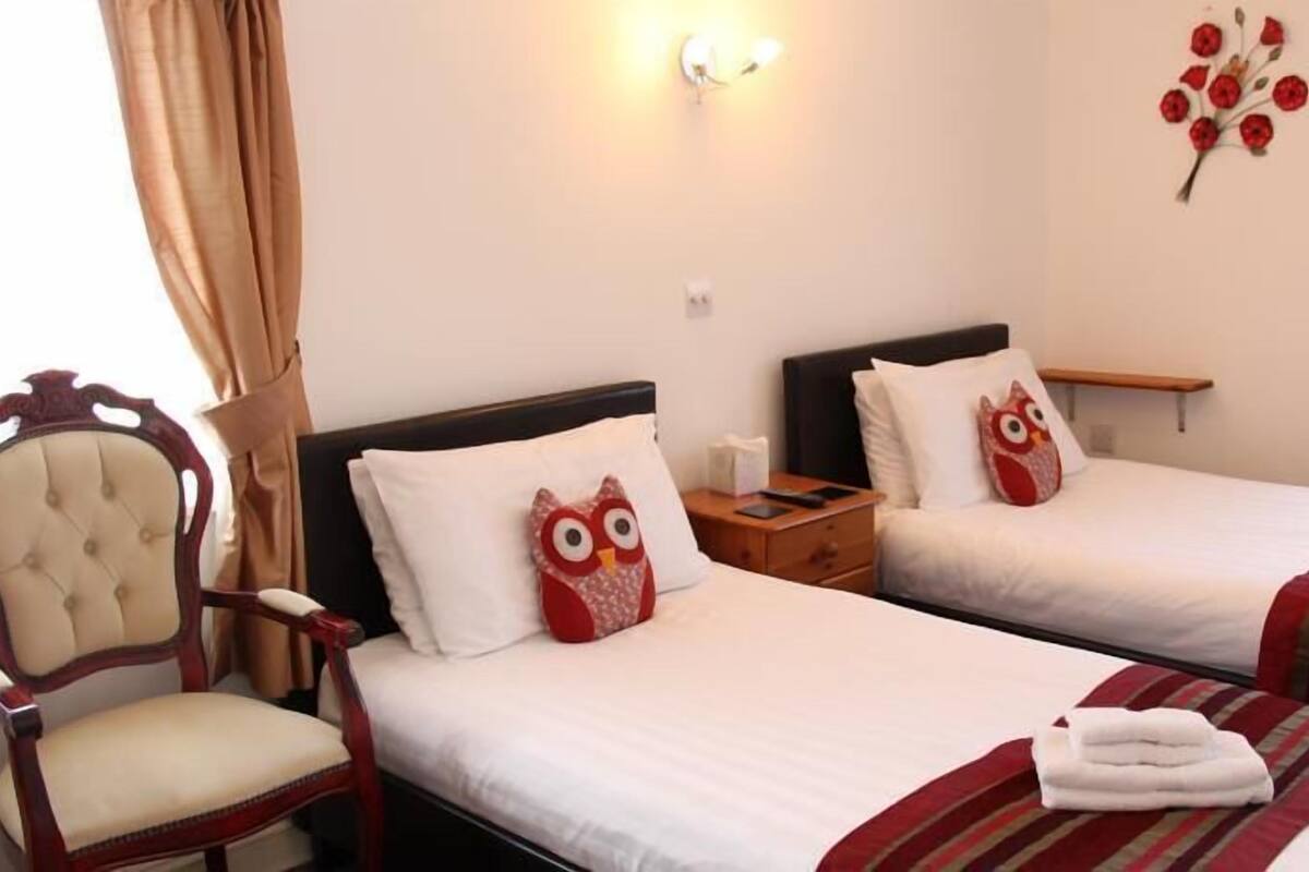A hotel room with two beds, each with an owl pillow, a wooden nightstand, a chair, and a wall-mounted light fixture.