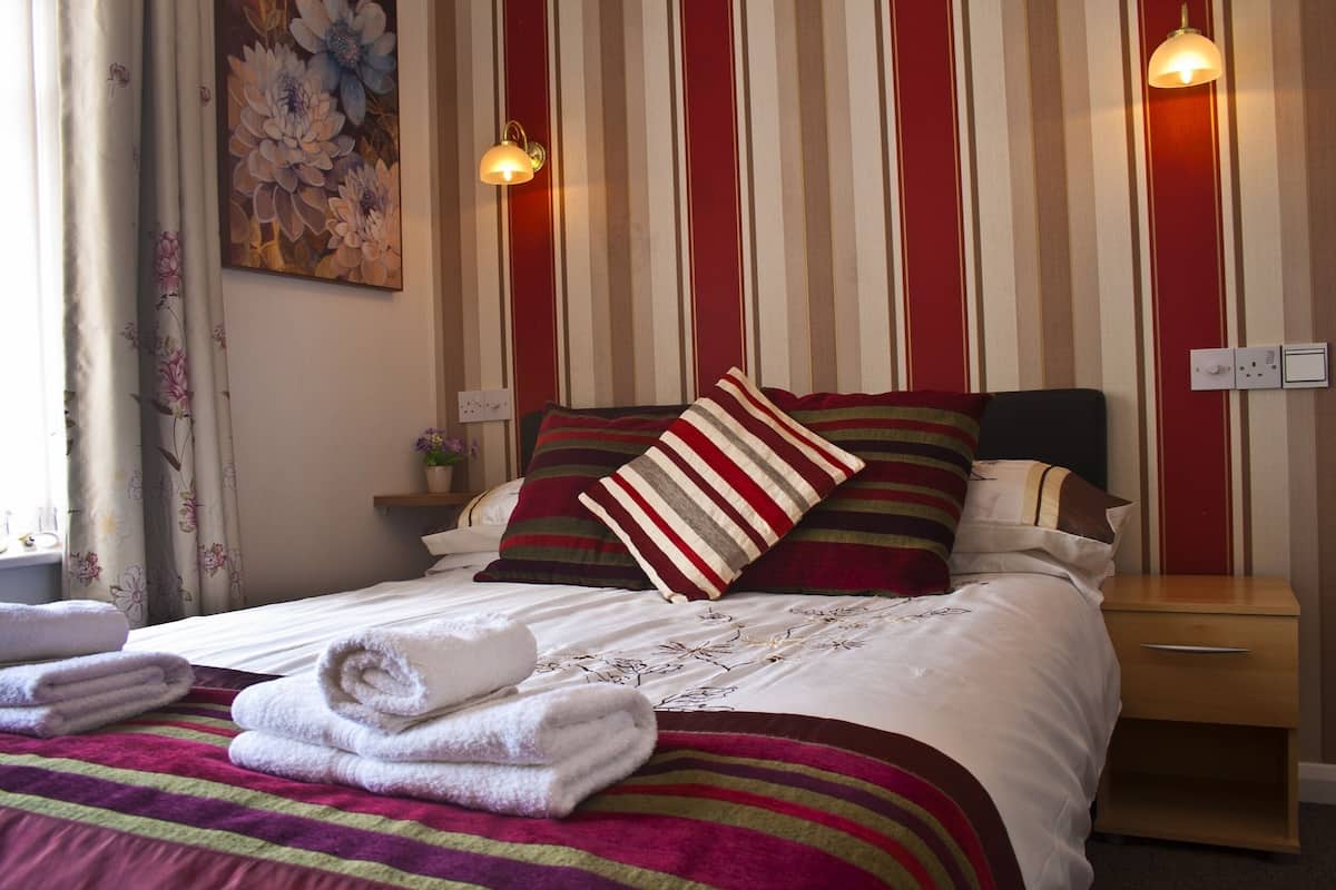 A neatly made bed with striped bedding and pillows, a bedside table with a lamp, and a floral painting on the wall.
