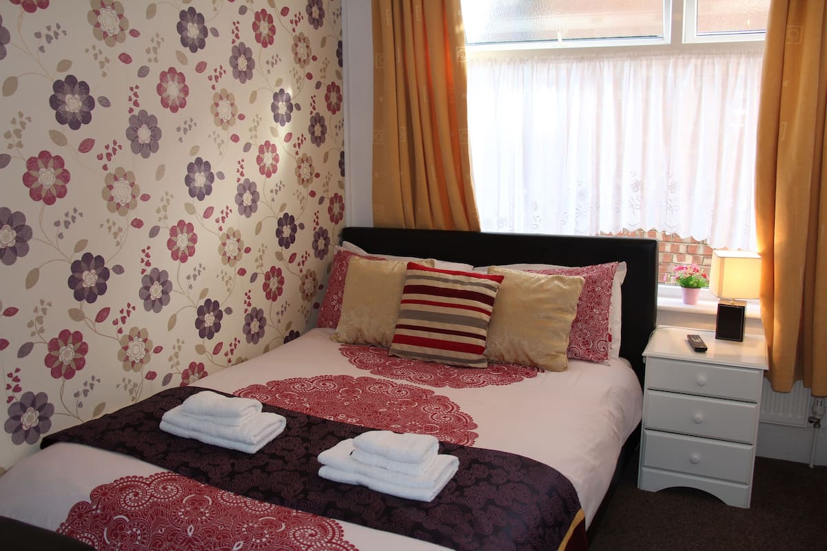 A neatly made bed with a floral-patterned wall, a bedside table with a lamp, and a window with curtains.