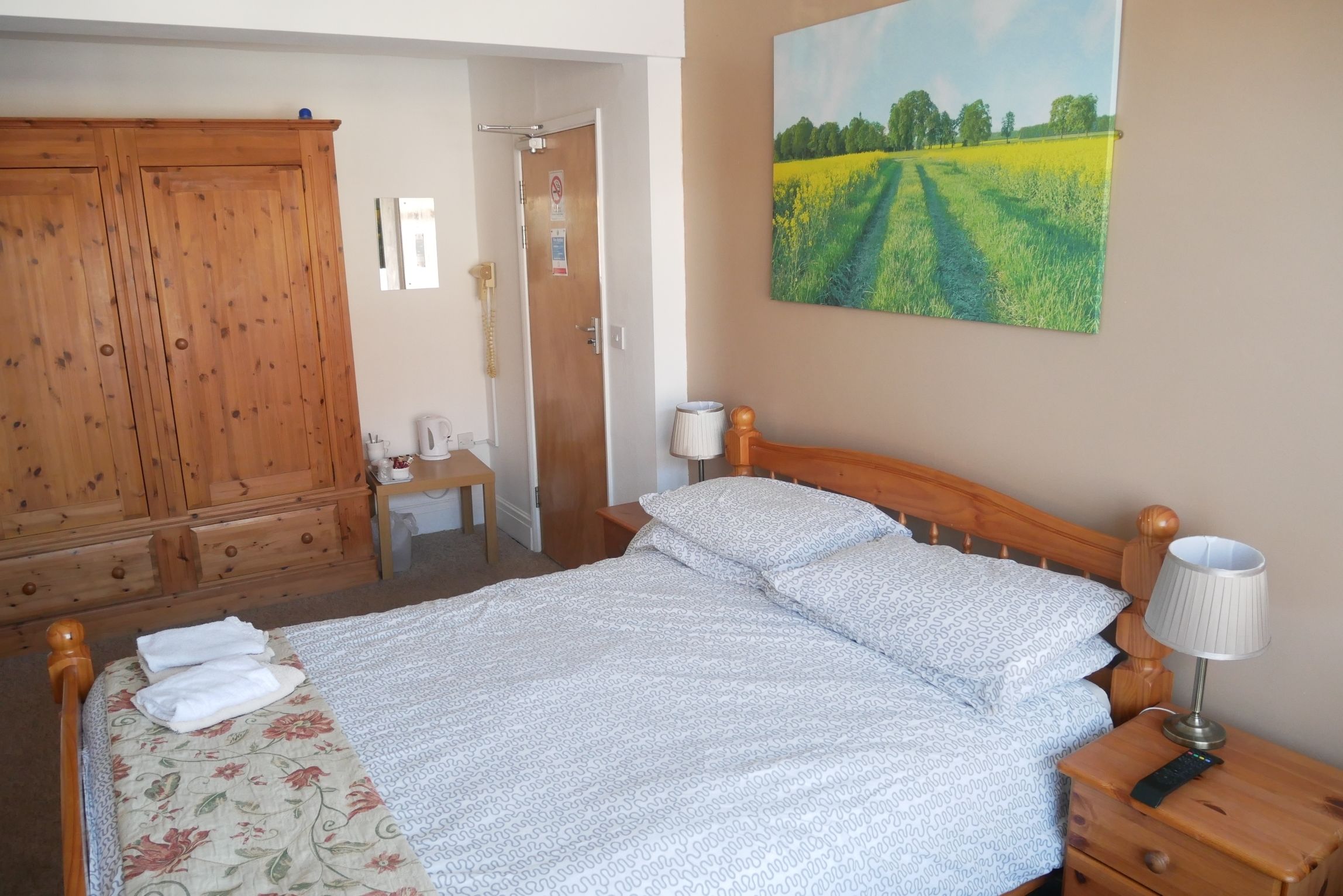 A bedroom with a bed, wooden wardrobe, a desk with a chair, a lamp, and a painting of a field.