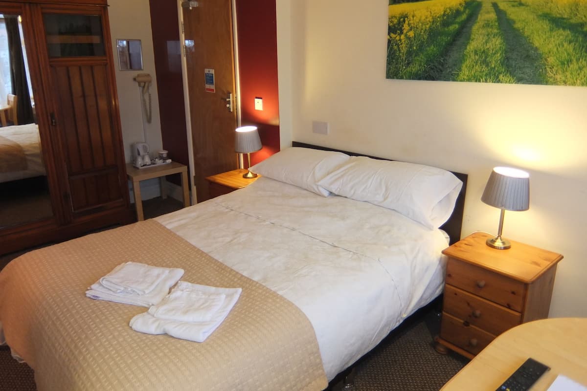 Habitación de hotel con una cama, dos lámparas de noche, una mesita de noche de madera, un armario de madera y un cuadro de un campo en la pared.