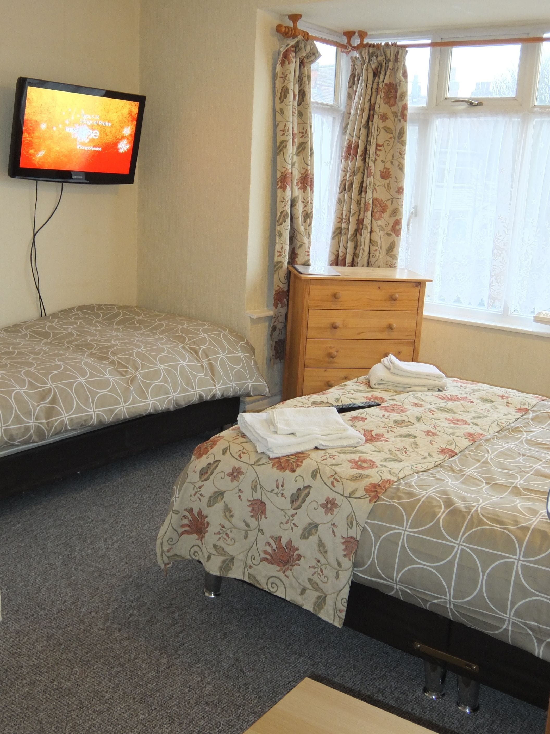 A bedroom with two beds, a wooden dresser, a television, and a window with floral curtains.