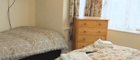 A bedroom with two beds, a wooden dresser, a television, and a window with floral curtains.