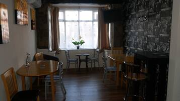 Uma área de refeições com mesas e cadeiras de madeira, uma pequena mesa redonda com uma planta em vaso, e um bar com banquetas.