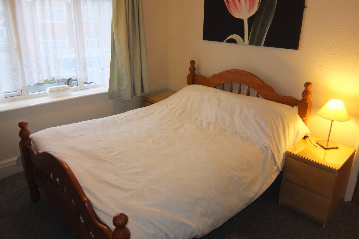 Una cama bien hecha con cabecera de madera, una mesita de noche con lámpara y una ventana con cortinas.