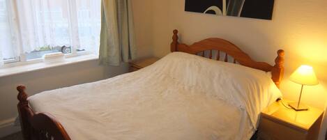 A neatly made bed with a wooden headboard, a bedside table with a lamp, and a window with curtains.