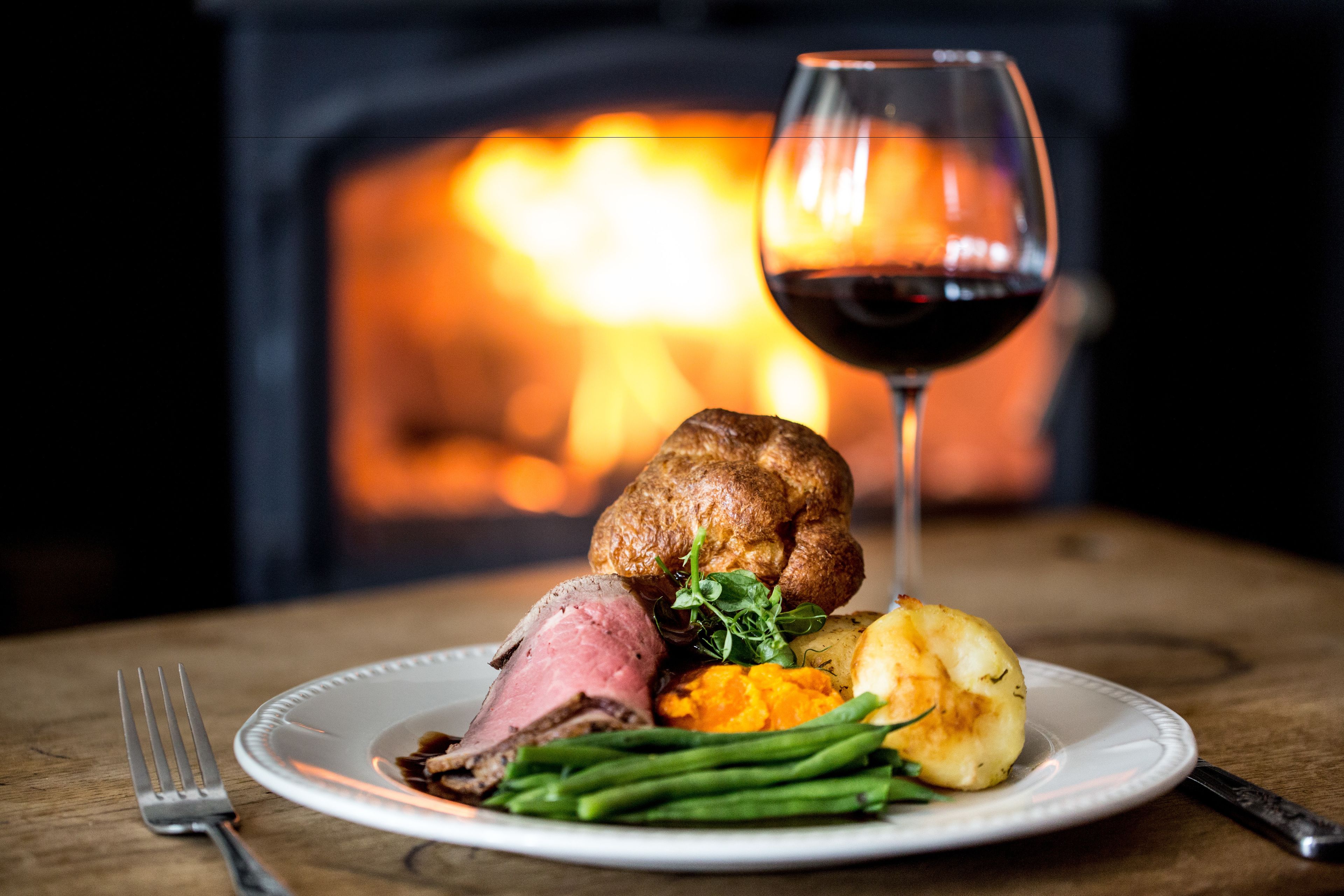 Un piatto di cibo con arrosto di manzo, Yorkshire pudding, fagiolini e purè di patate, accompagnato da un bicchiere di vino rosso, su uno sfondo con un camino.