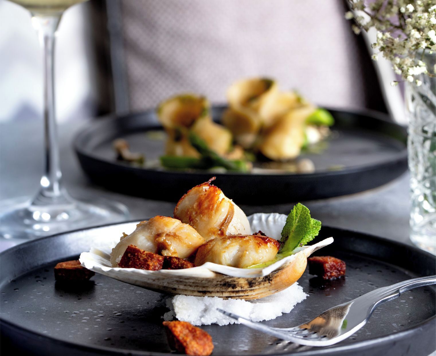 A plate of scallops with a garnish, accompanied by a glass of white wine and a vase of flowers.