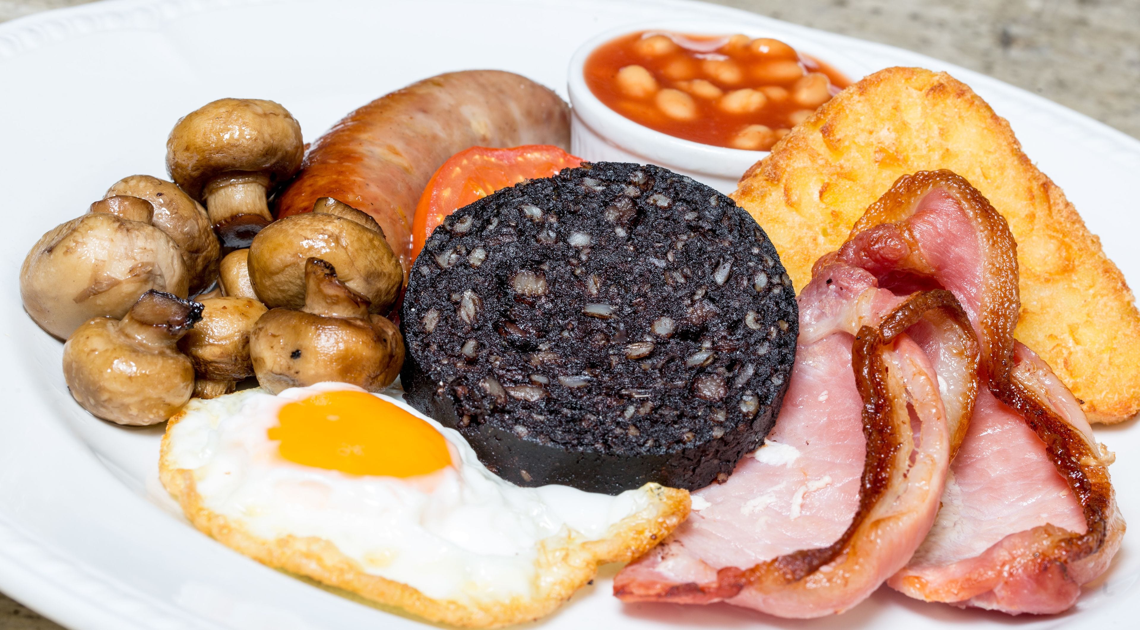 A plate of breakfast food including eggs, bacon, sausages, mushrooms, black pudding, baked beans, and toast.