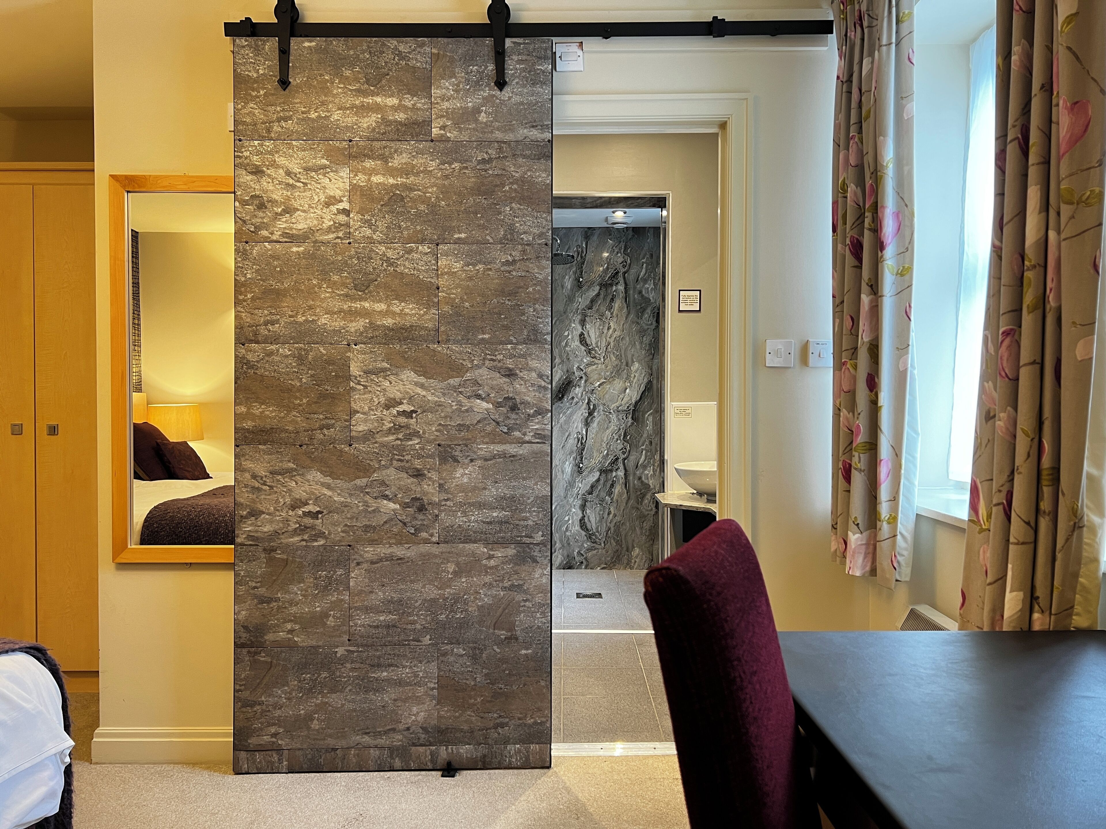 A room with a stone wall partition, a bed with a headboard, a mirror, a bathroom with a sink, and a window with floral curtains.