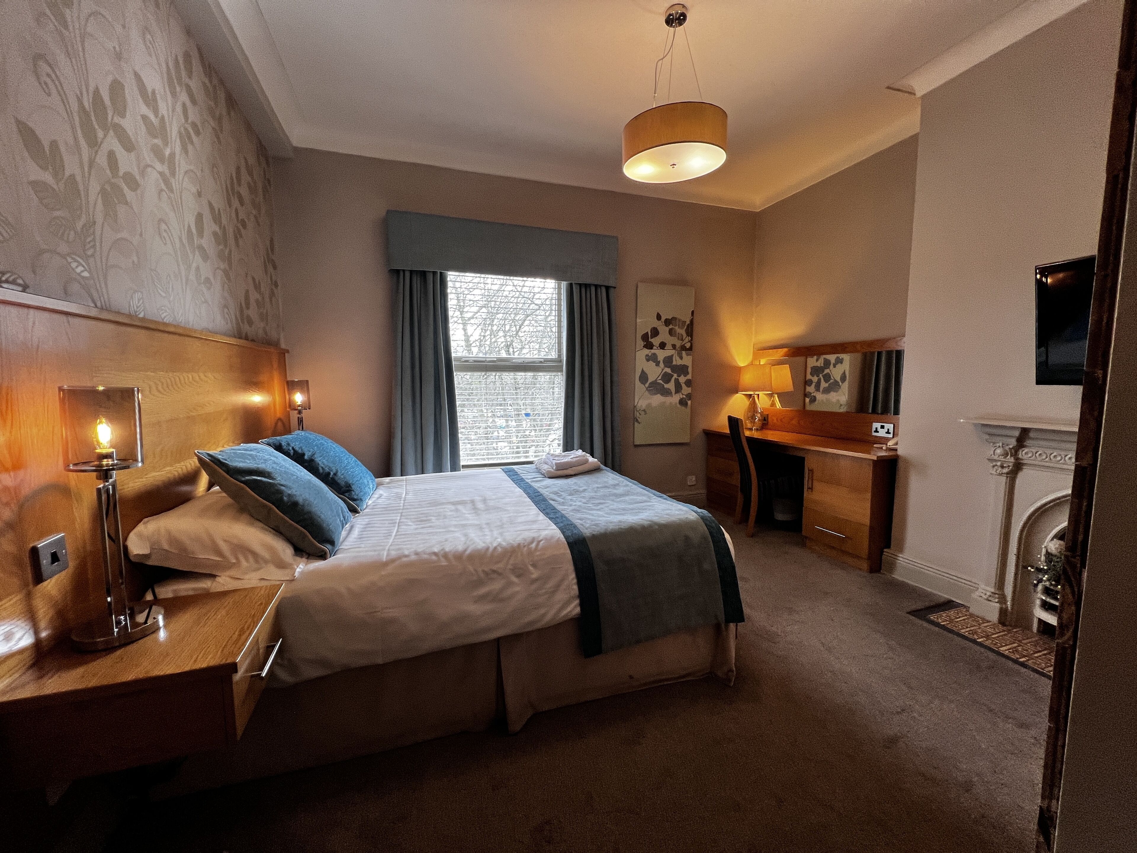 A hotel room with a bed, bedside table, lamp, desk, chair, and a fireplace.