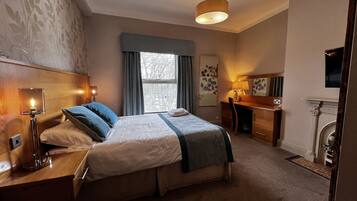 A hotel room with a bed, bedside table, lamp, desk, chair, and a fireplace.