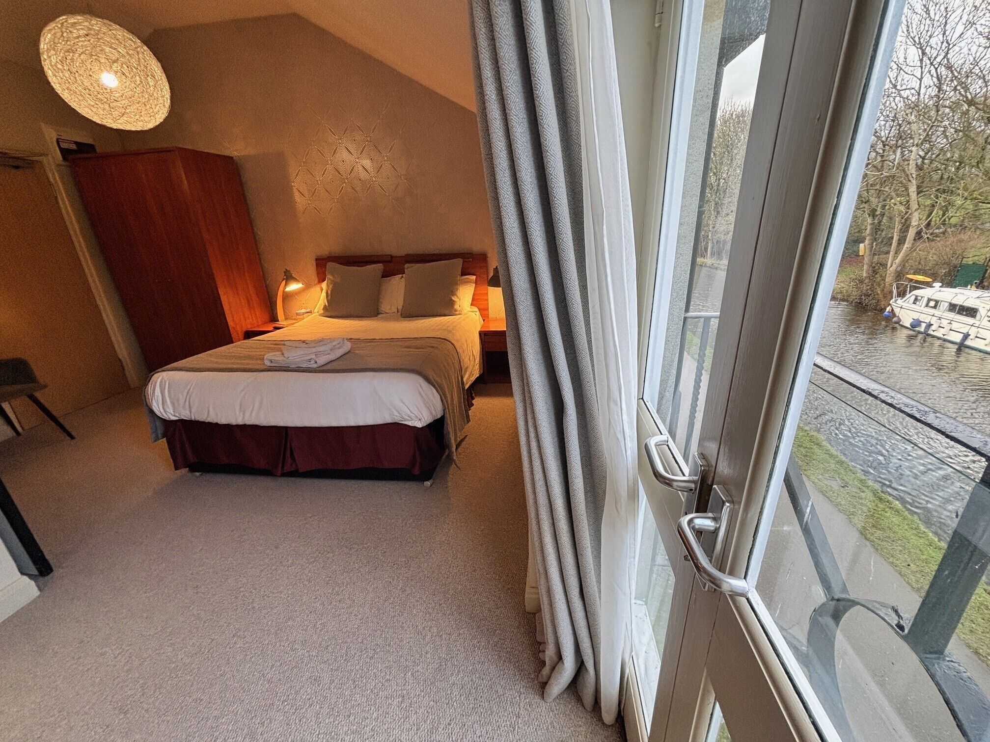 A bedroom with a large bed, a bedside table with a lamp, and a view of a canal outside.