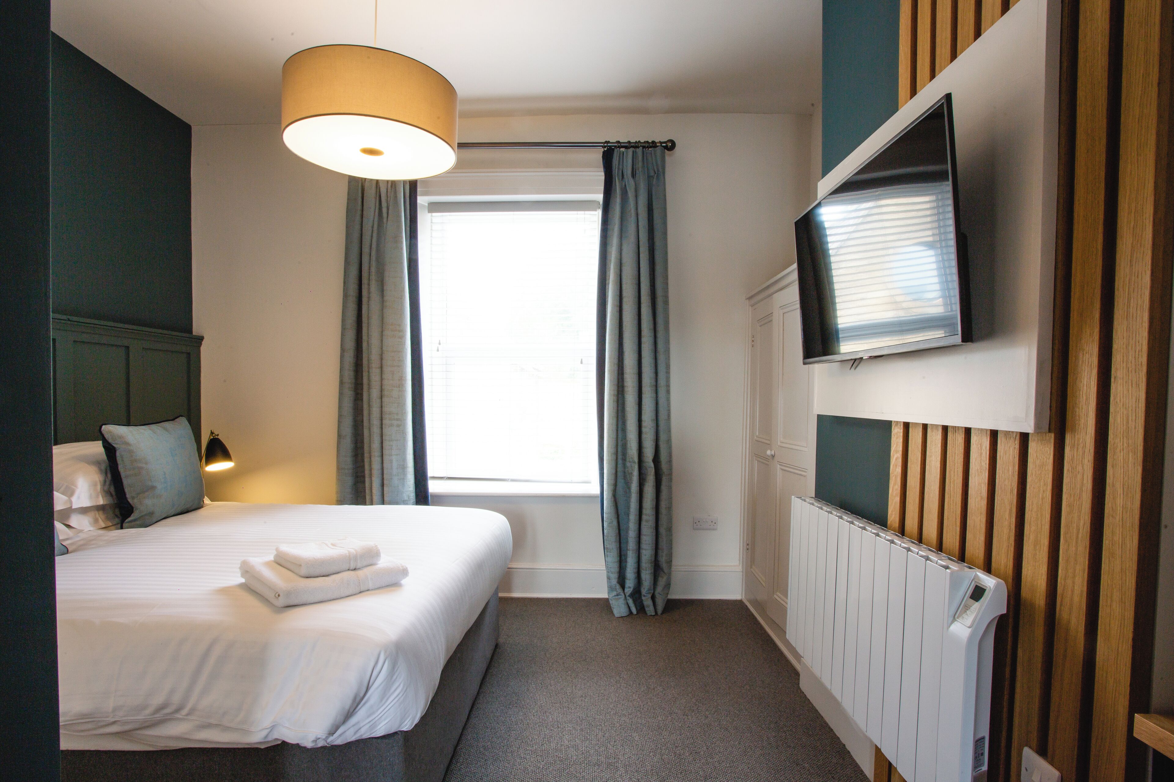 Una habitación de hotel con una cama, un televisor montado en la pared, una ventana con cortinas y un radiador.