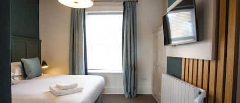 A hotel room with a bed, a TV mounted on the wall, a window with curtains, and a radiator.