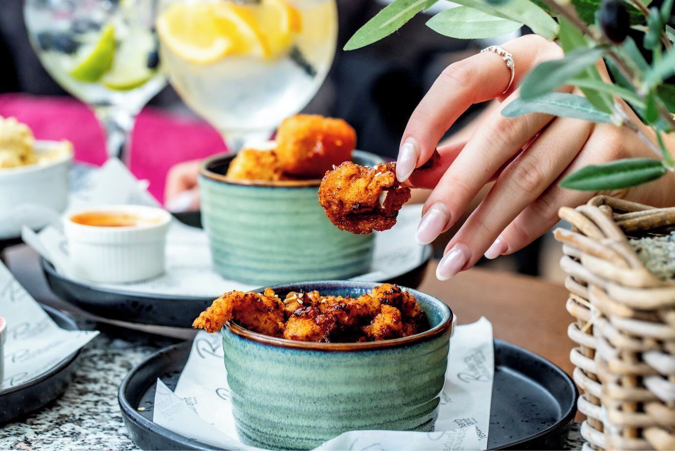 A person holding a piece of fried chicken, with bowls of dipping sauce and a drink in the background.