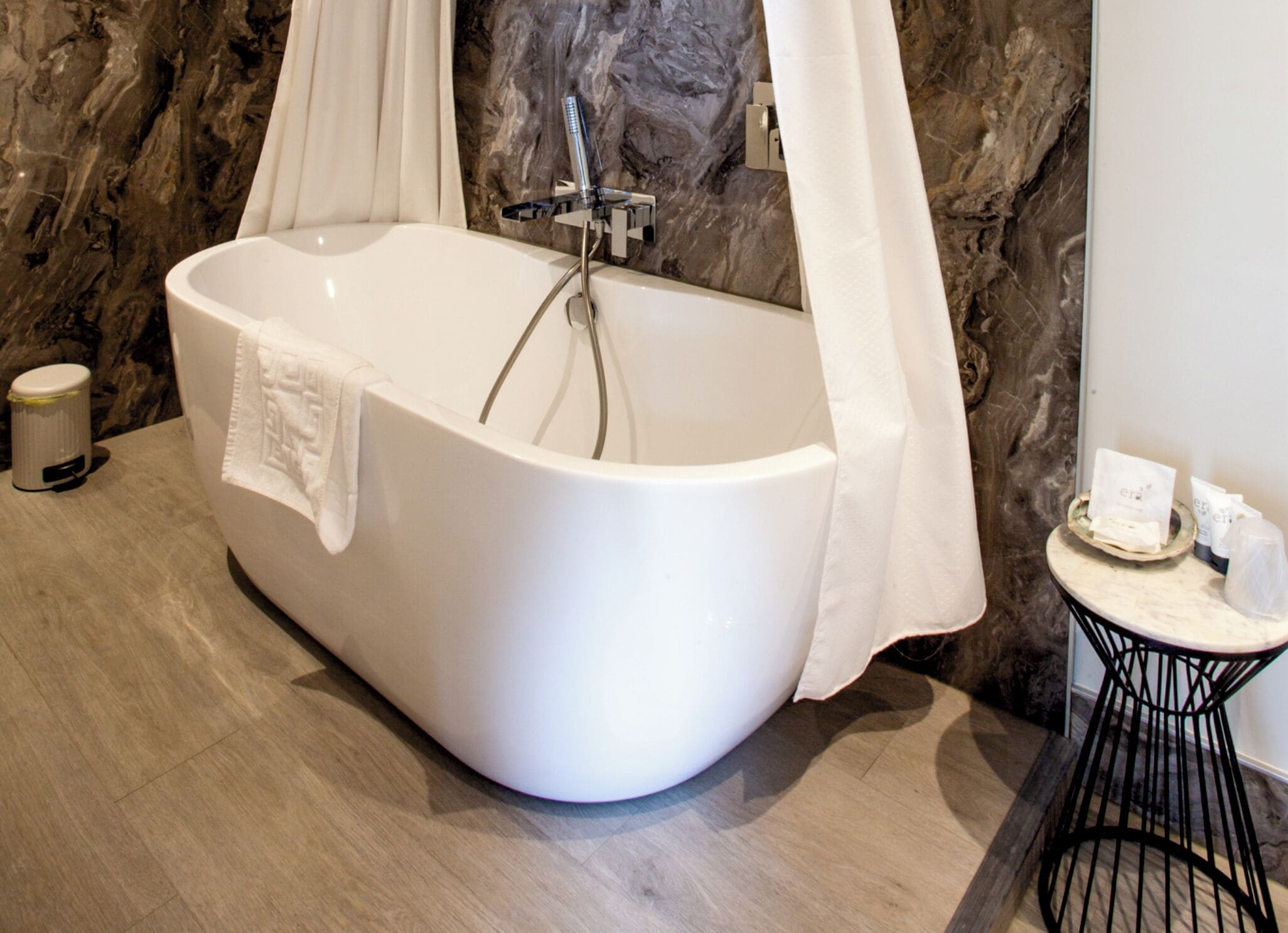 Een moderne badkamer met een vrijstaand bad, marmeren wanden, een klein rond tafeltje met toiletartikelen en een afvalbak.