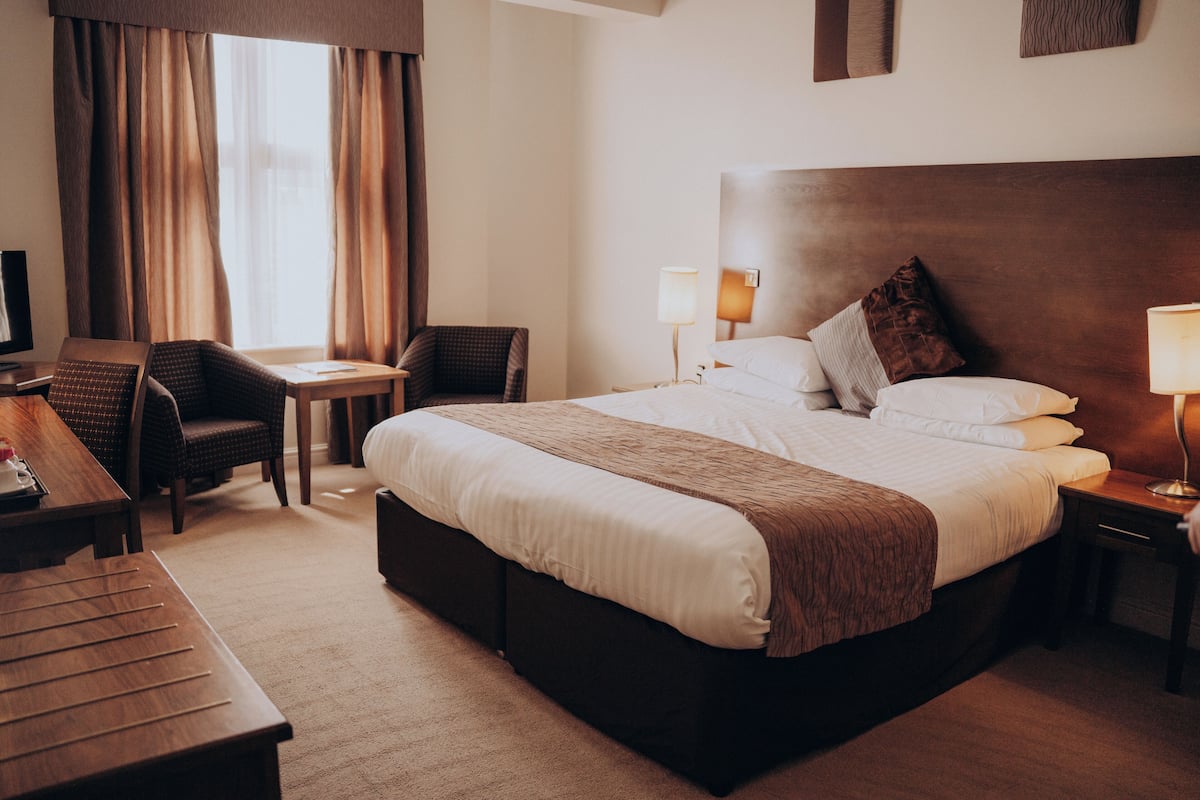 A hotel room with a large bed, a desk, a chair, a bedside table with a lamp, and a television.