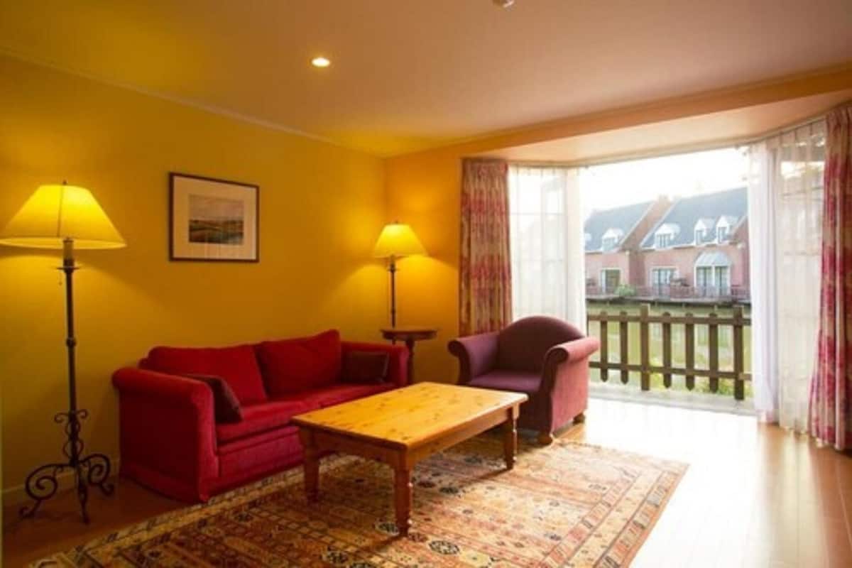 Habitación con un sofá rojo, una mesa de madera, dos lámparas de pie, un tapete estampado y vista a una casa a través de una puerta de vidrio.