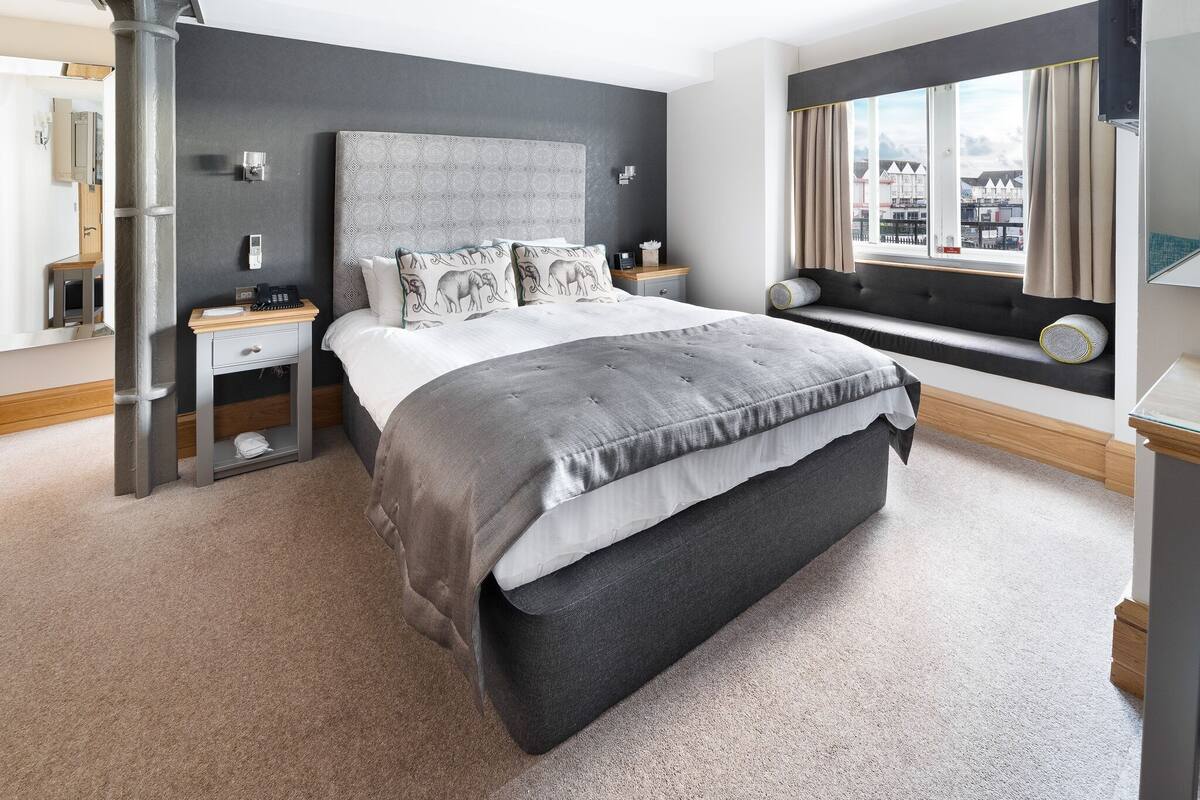 Un dormitorio moderno con una cama grande, una mesita de noche, un asiento junto a la ventana y vistas a la ciudad.
